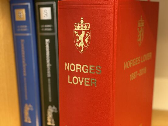 3 bøker i ei bokhylle. Den fremste, som er i fokus, er tjukk med raud perm og teksten "Norges lover" i gullfarga skrft.