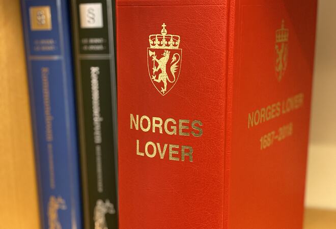 3 bøker i ei bokhylle. Den fremste, som er i fokus, er tjukk med raud perm og teksten "Norges lover" i gullfarga skrft.