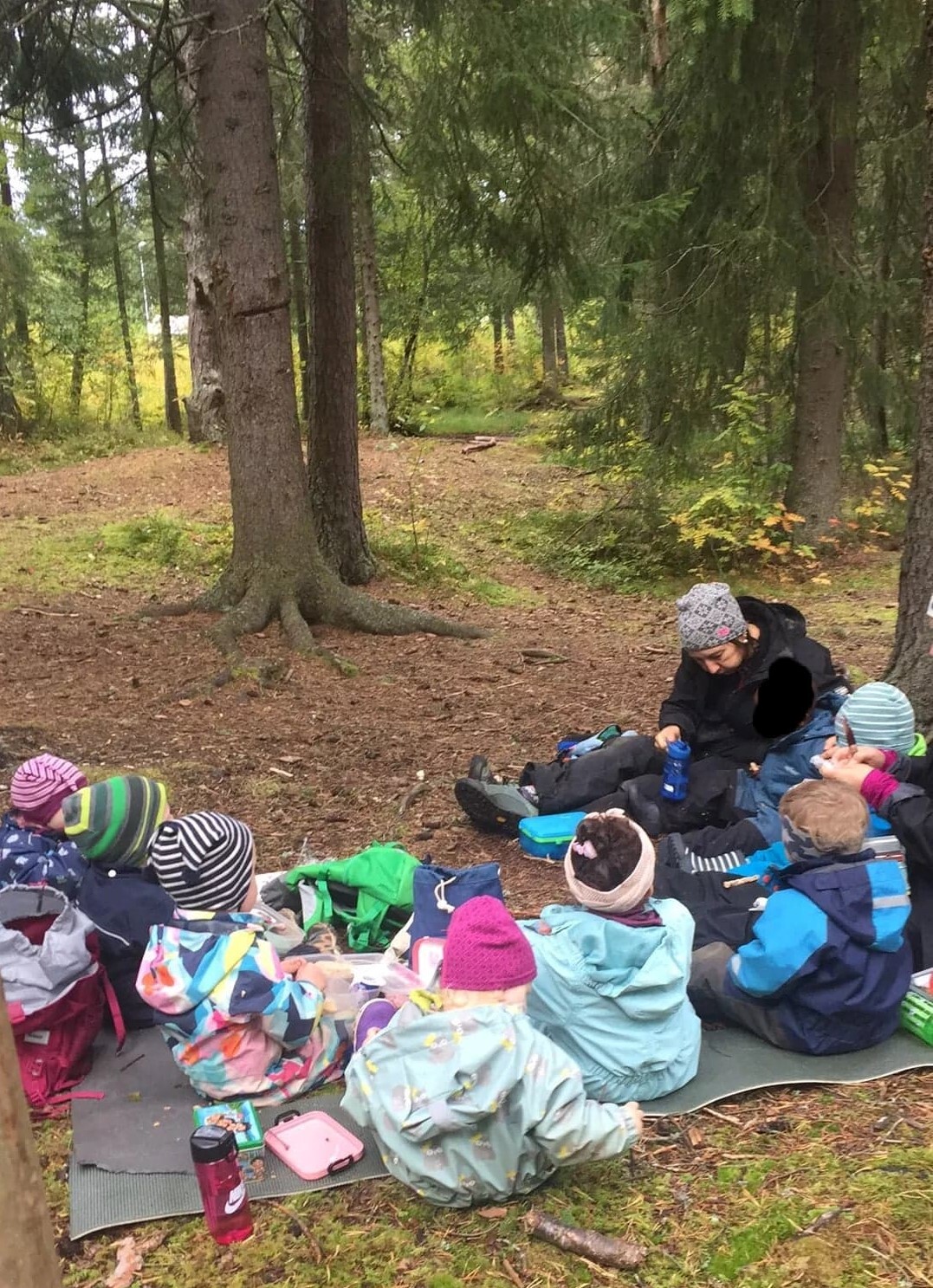 Barn og en voksen sitter sammen i en halvsirkel i skogen, og spiser lunsj.