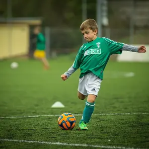 Bildet viser en liten gutt på en fotballbane, kledd i hvit og grønn fotballdrakt, som akkurat skal til å sparke en fotball.