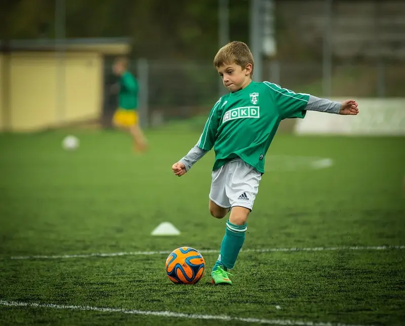Bildet viser en liten gutt på en fotballbane, kledd i hvit og grønn fotballdrakt, som akkurat skal til å sparke en fotball.