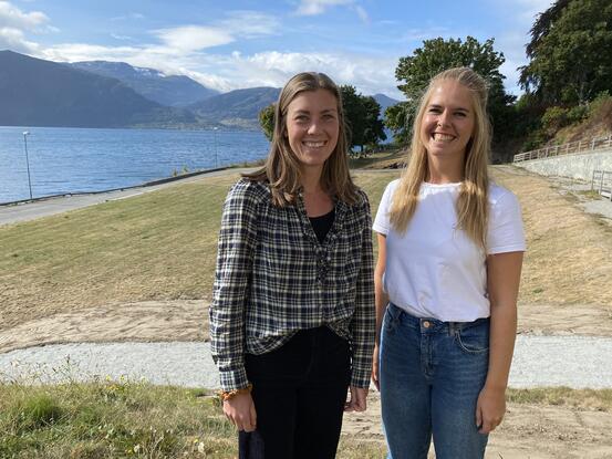 To jenter som smiler til kameraet, ei i rutetet skjorte og ei i kvit t-skjorte. Fjord og fjell i bakgrunnen.