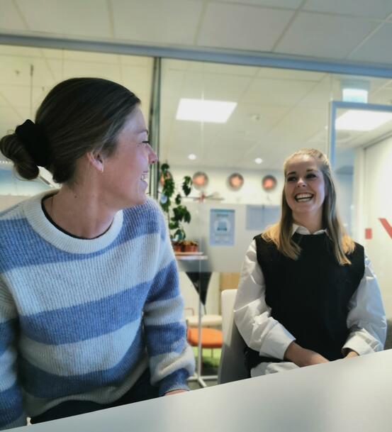 To personar ved ei bord i kontorlandskap, smiler mot kvarandre.