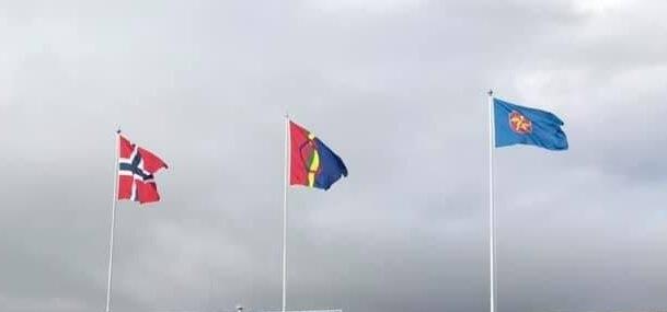 Bilde av norsk, samisk og kvensk flagg på hver sin flaggstang mot skyet himmel