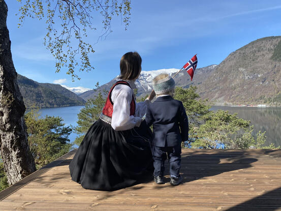 Dame i bunad og liten gut i dress med flagg, utsyn over fjord og fjell