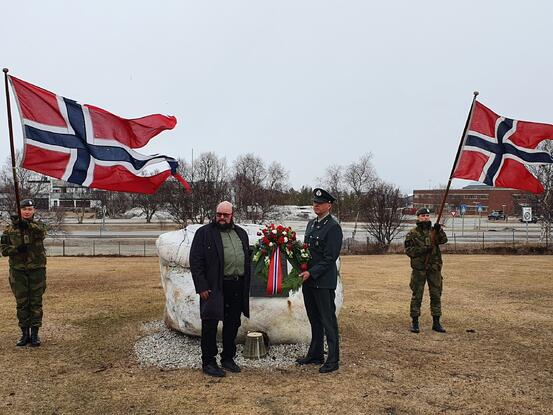 To soldater med norske flagg står på hver side av to menn som legger ned krans ved et monument