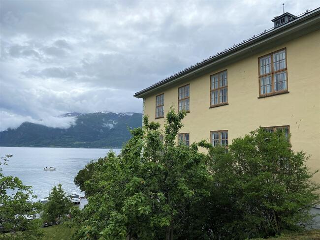 Gul murbygning med grønebusker framfor. Liten kvit båt på fjorden.