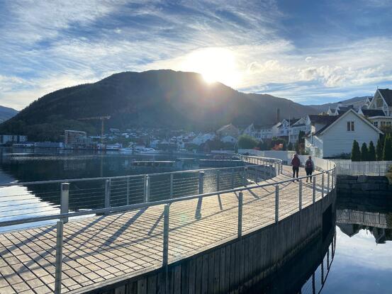 To personar som går tur på fjordstien i Sogndal. Fjord, fjell og motlys frå kveldssola.