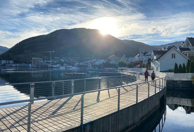 To personar som går tur på fjordstien i Sogndal. Fjord, fjell og motlys frå kveldssola.