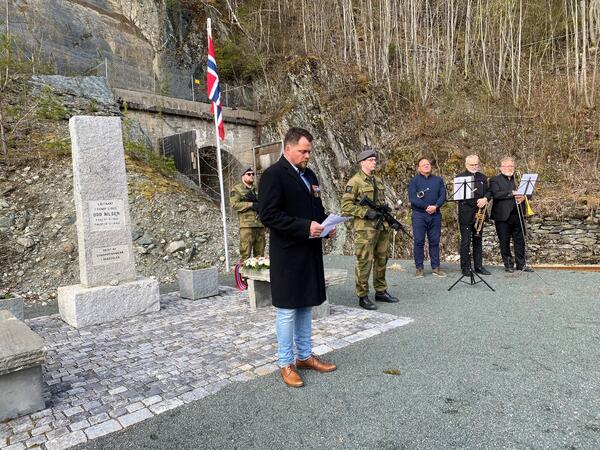 Mann i sort jakke holder tale, mens militærkledde menn holder vakt. Tre mannlige musikere i bakgrunnen.