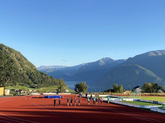 Ei gruppe ungdommar joggar på friidrettsanlegg, sol, fjord og fjell.