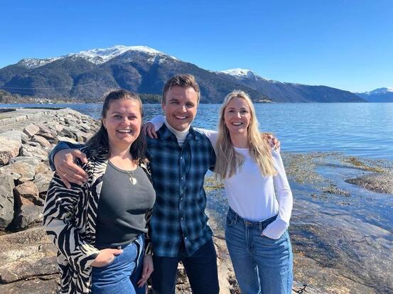 Tre personar som held rundt kvarandre og smiler inn i kameraet. Fjord og fjell i bakgrunnen.