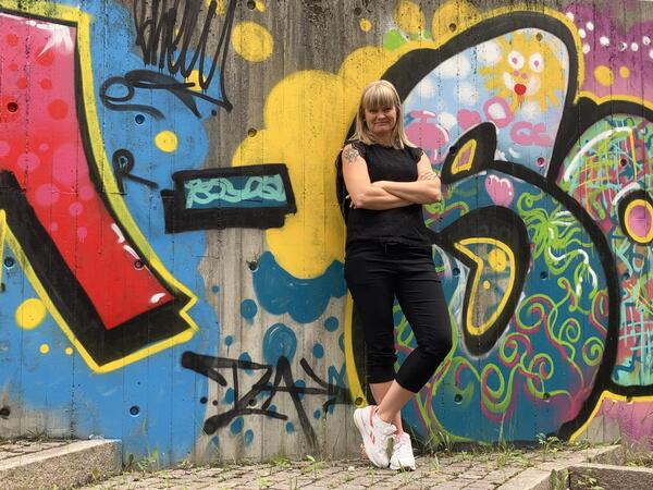 Dame i svarte kle med lyst hår står framfor ein fargerik grafittivegg.