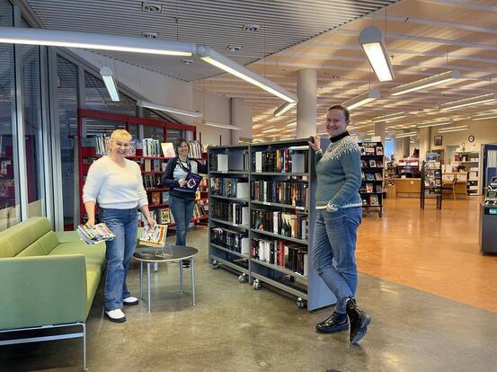 Sofa, bord og bibliotekshyller med bøker. Tre personar ser smilande inn i kameraet.