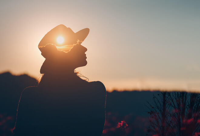 Silhuetten av en person med cowboyhatt mot en oransje solnedgang. Personen står i profil og solen er plassert perfekt i midten av hatten, noe som skaper en illusjon av at solen er en del av hatten. Bakgrunnen viser en svakt opplyst himmel med farger