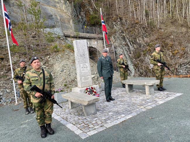 Minneseremoni ved et utendørs minnesmerke. Det er flere personer i militæruniform til stede, noen bærer våpen. Det er også norske flagg på stedet. Seremonien ser alvorlig og formell ut, med deltakerne i oppmerksom holdning.