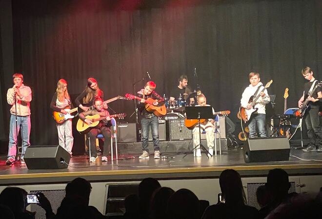 En gruppe unge musikere opptrer på en scene. Det er flere gitarister, en trommeslager, en keyboardist, og en sanger. Konsert eller en skoleforestilling.