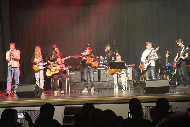 En gruppe unge musikere opptrer på en scene. Det er flere gitarister, en trommeslager, en keyboardist, og en sanger. Konsert eller en skoleforestilling.