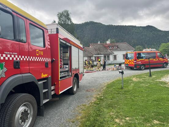 En nødssituasjon hvor flere brannbiler er utplassert. Det er en stor rød brannbil i forgrunnen, og en mindre rød utrykningsbil til høyre i bildet. Bak brannbilene kan man se noen brannmenn.