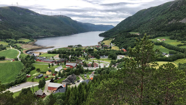 På bildet ser du et idyllisk tettsted ved en fjord, omringet av frodige grønne åser og fjell. Landskapet er preget av velholdte gårder, hus og en vindmøllepark på fjellet i bakgrunnen.