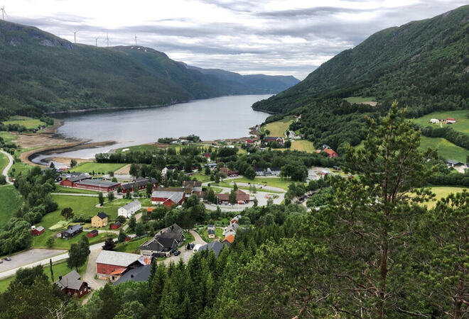 På bildet ser du et idyllisk tettsted ved en fjord, omringet av frodige grønne åser og fjell. Landskapet er preget av velholdte gårder, hus og en vindmøllepark på fjellet i bakgrunnen.