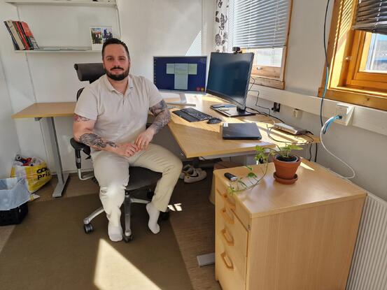 Bilde av mann (Nemanja Radisevic) som sitter på kontorstol framfor bord med pc skjermer.