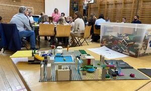 Legobygg i framgrunnen, vaksne og ungar på bord i bakgrunnen