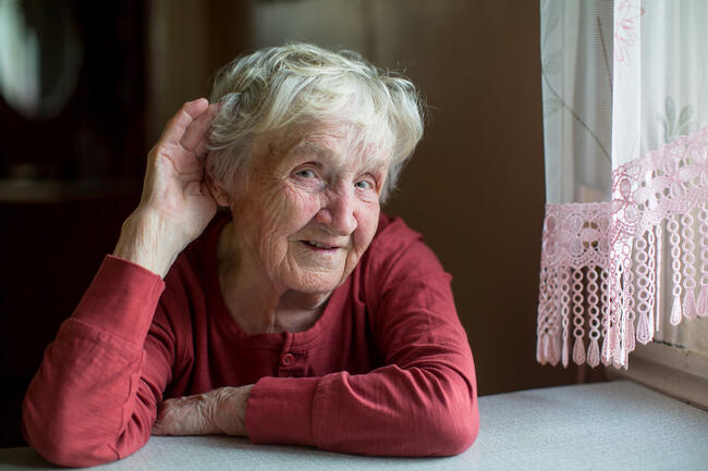 En eldre kvinne med hvitt hår sitter ved et bord. Hun har på seg en rød genser og støtter hodet med hånden, mens hun holder den andre hånden bak øret, som om hun prøver å høre bedre.