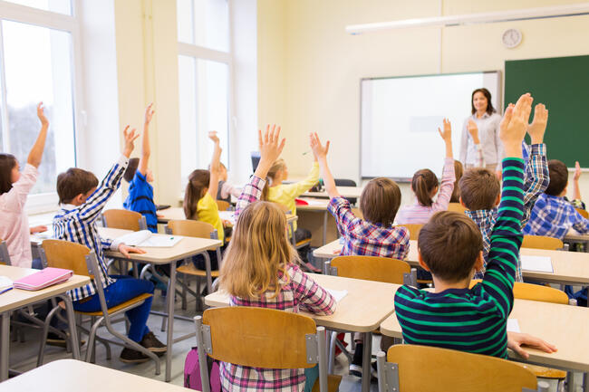 Skoleelever i et klasserom med læreren i bakgrunnet. Elevene sitter med ryggen til.