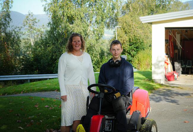 Bilde av en kvinne og en ung mann. Kvinnen står ved siden av den unge mannen, som sitter på en gressklipper med hodetelefoner rundt halsen. I bakgrunnen er det et uthus og trær, med et fjellandskap i det fjerne. Kvinnen har på seg en lys kjole og en hvit cardigan, mens mannen har en mørk hettegenser og arbeidsbukser.