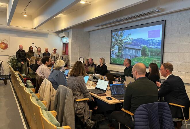 Seks personar (representantar for råda) står ved enden av eit møtebord. Finansutvalsmedlemmene sit rundt bordet.