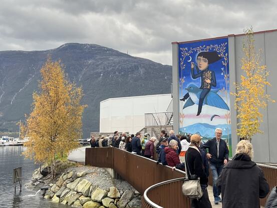 Bilete frå gangbrua over Sogndalselvi mot Norturaveggen med utsmykkinga. Ein person på ein blå fugl teiknar månen. Under er det eit fuglebur.