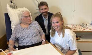 Eldre kvinne med kort, grått hår sit på eit pasientrom på sjukeheimen saman med ein dresskledd mann og ei kung kvinne i kvit uniform