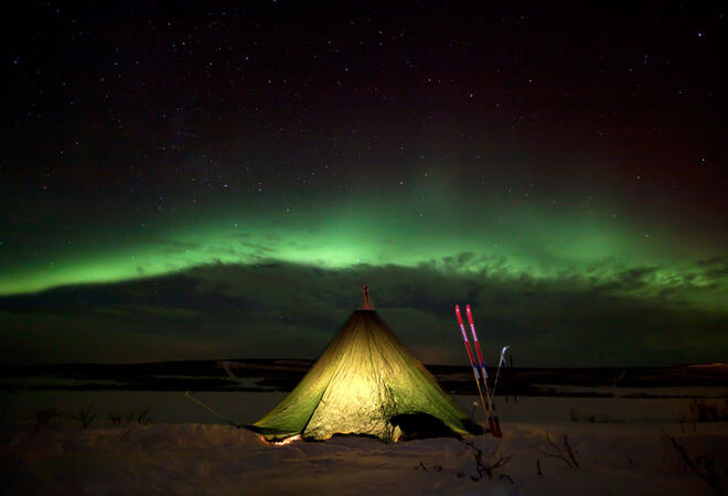 Grønn lavvu opplyst innenfra under nordlyset. Det er vinter, måneskinn og kaldt.