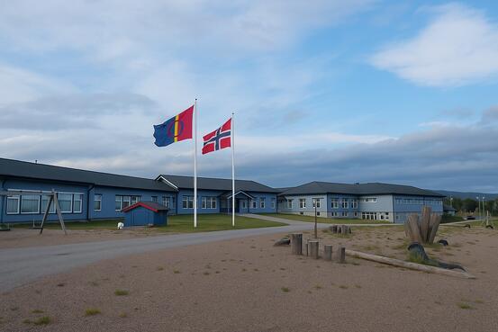 Nesseby oppvekstenter. Foto av Nesseby oppvekstenter. Et blått bygg, blå himmel og det samiske og norske flagget vaiende i forgrunnen.