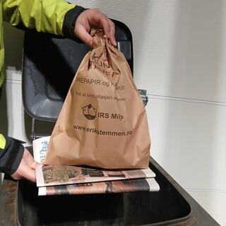 Bilde av to hender som holder en avis under en pose med matavfall, over en avfallsbeholder.