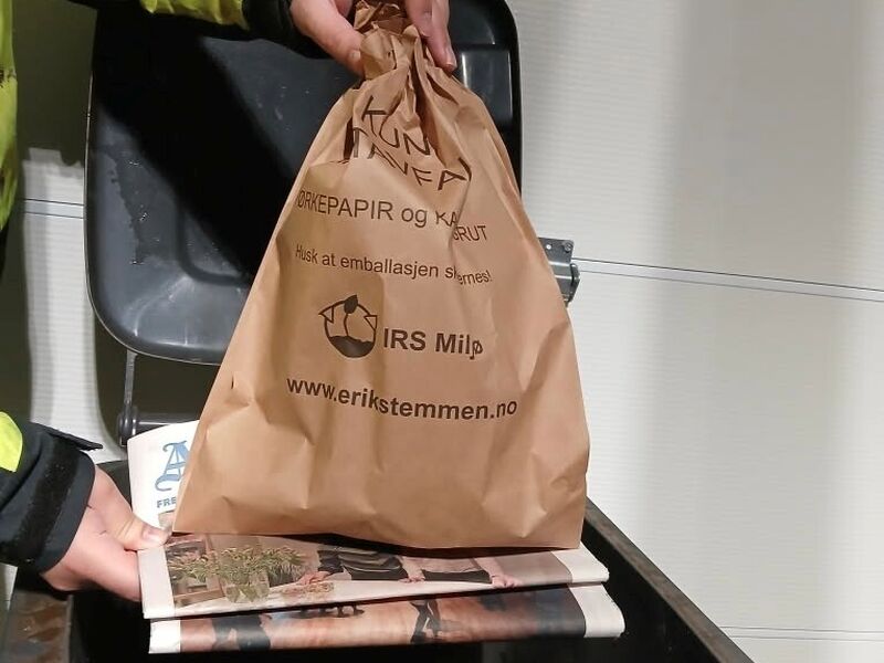 Bilde av to hender som holder en avis under en pose med matavfall, over en avfallsbeholder.